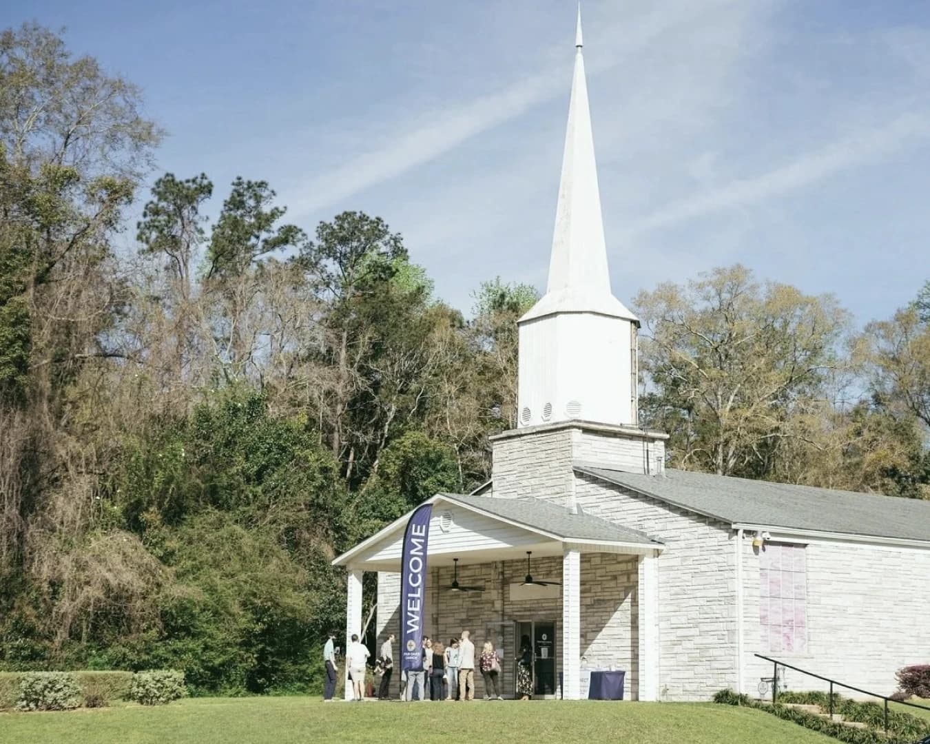 A white church with a tall steeple, people gathering under a Welcome banner at the front doors