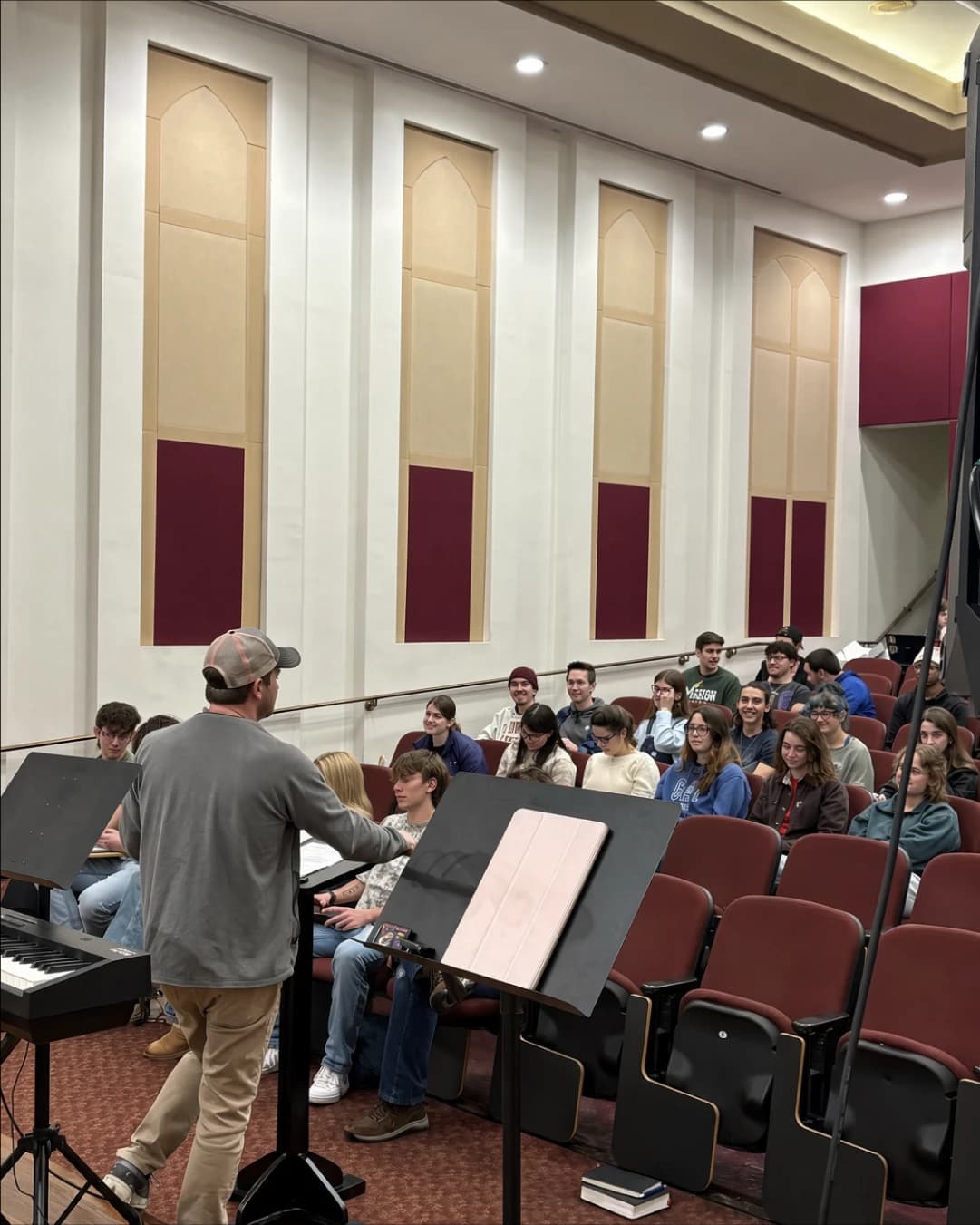 A worship leader at a keyboard facing rows of students in the auditorium seats at Reach Night