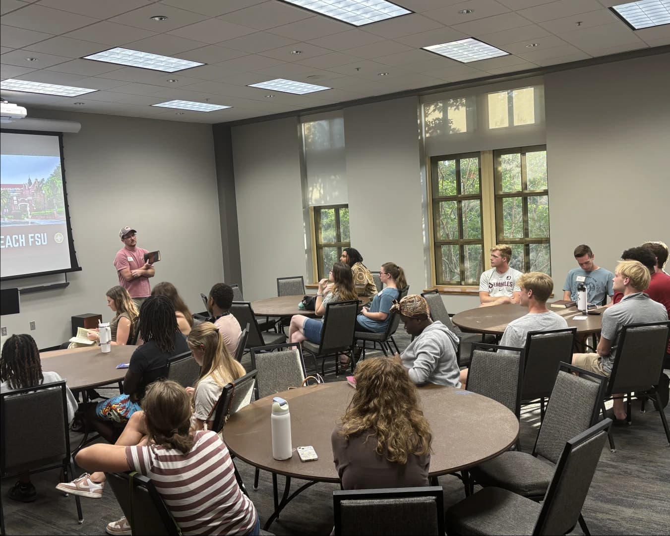 Students seated around round tables in a campus meeting room, listening to someone teaching at the front